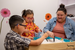 Mother and two young children playing with toys in a playroom at a sandbox table.