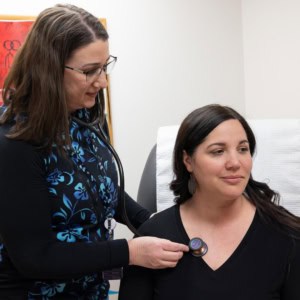 Female provider offering care to female patient by checking heart rate with a stethoscope in a clinical setting.