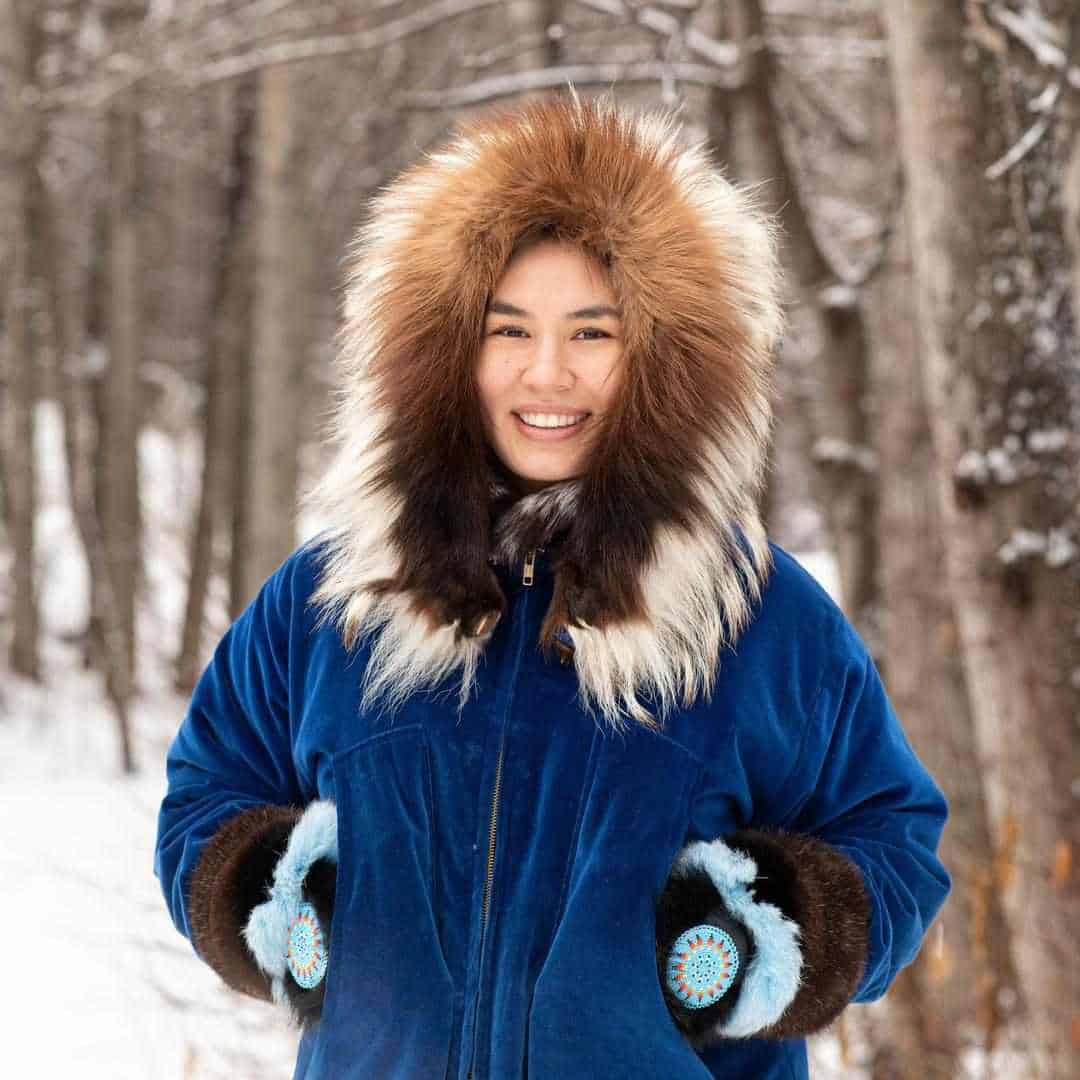 Alaska Native Woman in traditional regalia of a fur-trimmed parka standing in a snowy, wooded scene.